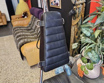 May include: Black leather motorcycle backrest with a padded, rectangular design. The backrest is mounted on a chrome support, attached to a black motorcycle seat. The backrest has a diamond-shaped top.