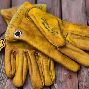 May include: A pair of yellow leather gloves with stitching detail. The gloves have a flap on the back of the hand with the word "Sharoq" and "Thailand" printed on it. The gloves are made of genuine leather.