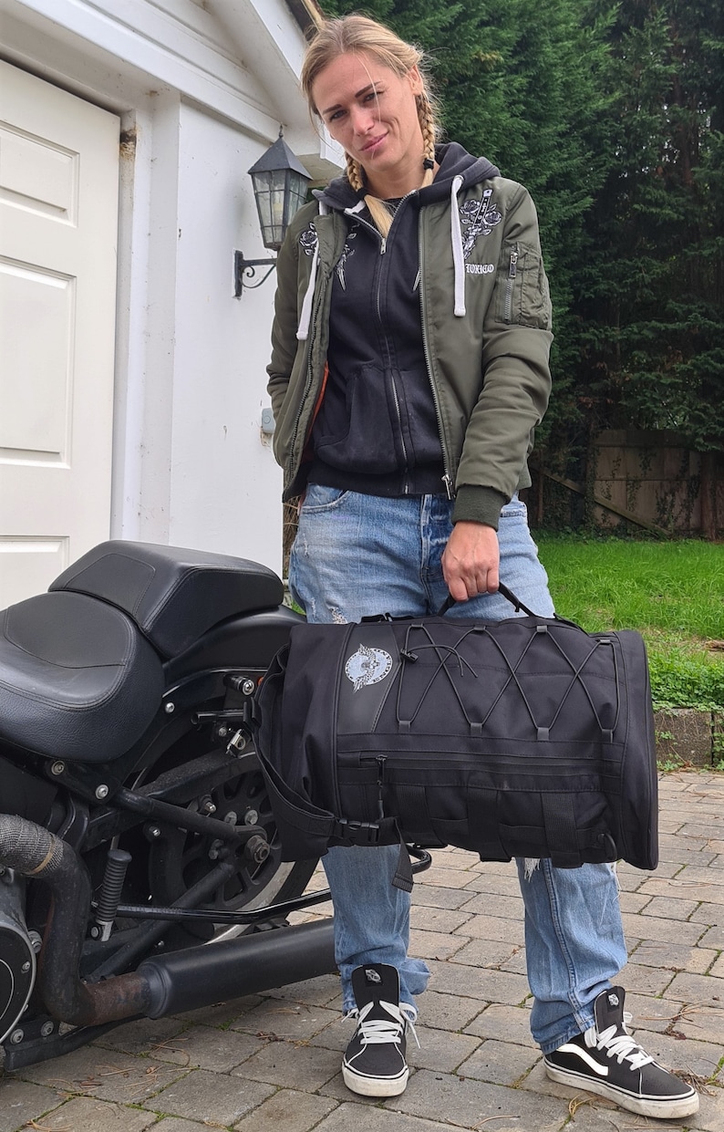 May include: A woman wearing a green jacket with a black hoodie underneath, blue jeans, and black sneakers, is standing next to a black motorcycle. She is holding a black motorcycle bag with a white logo.
