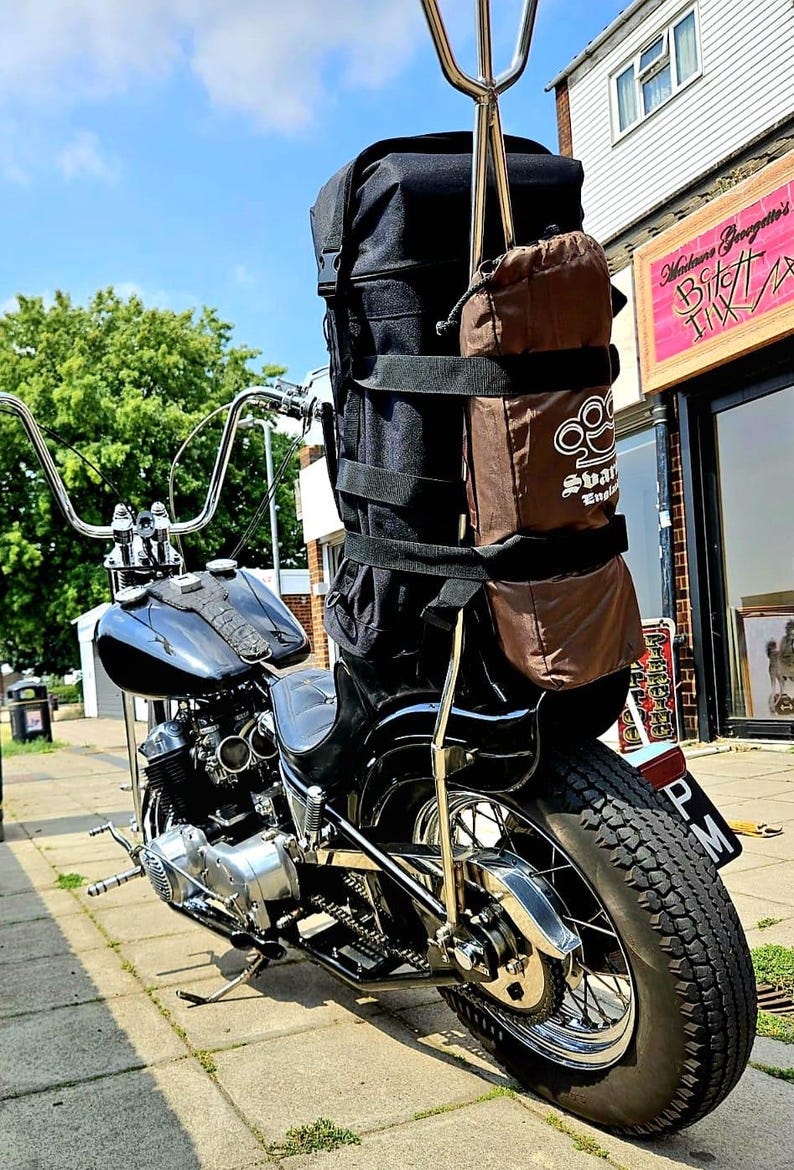 May include: A black motorcycle with a brown and black saddlebag attached to the back. The motorcycle has a chrome exhaust pipe and a chrome rear wheel. The motorcycle is parked on a sidewalk in front of a building.