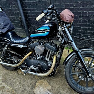 May include: Black Harley Davidson motorcycle with blue and white stripes on the gas tank. The motorcycle has a brown leather bag on the handlebars and a black bag on the back seat.