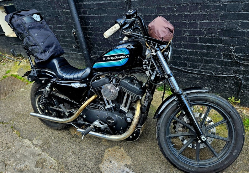 May include: Black Harley Davidson motorcycle with blue and white stripes on the gas tank. The motorcycle has a brown leather bag on the handlebars and a black bag on the back seat.