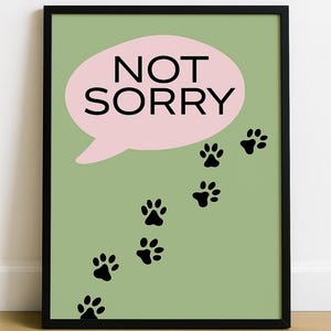May include: Framed art print with a pale green background. A pink speech bubble contains the words "NOT SORRY" in black. Black paw prints trail down the right side, suggesting a pet's path.