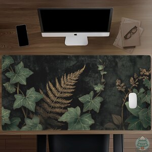 May include: A decorative desk mat featuring a dark green background with a gold fern and green ivy leaf design. A computer mouse rests on the mat, alongside a computer monitor, phone, and books. The mat is approximately 31.5 inches long and 11.8 inches wide.