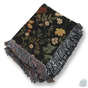May include: Folded black woven throw blanket with a floral pattern in shades of yellow, green, and brown. The blanket has a fringed edge with a mix of colours. The Cozy Spruce logo is in the bottom right corner.