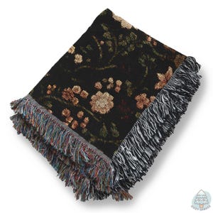 May include: Black woven throw blanket with a floral pattern in shades of tan, green, and red. The blanket has a fringed edge with a mix of colours. The blanket is folded and displayed on a white background.