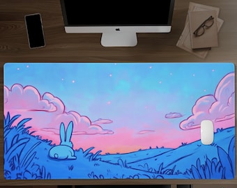 May include: A rectangular desk mat featuring a printed illustration of a blue rabbit sitting in a grassy field at sunset. The scene includes pink and blue clouds against a gradient sky with small glowing particles. A white computer mouse rests on the mat.