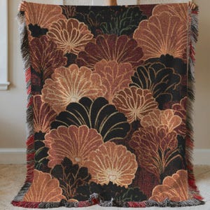 May include: A woven throw blanket featuring a repeating floral pattern in shades of brown, black, and gold. The design includes stylized flower shapes with detailed outlines. The blanket has a fringed edge in red and black.
