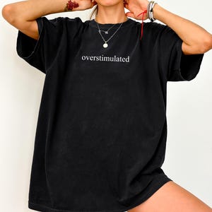 May include: Black oversized t-shirt with the white text "overstimulated". The shirt has short sleeves and a loose fit, ideal for casual wear. The fabric looks soft and slightly faded.