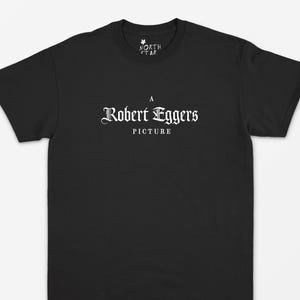 Op de afbeelding: Zwart t-shirt met witte tekst. De tekst bevat "NORTH STAR" erboven en "A Robert Eggers PICTURE" in een decoratief lettertype. Het shirt heeft een ronde hals.