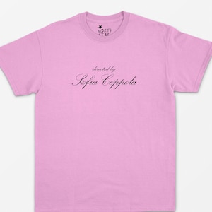 May include: A light pink t-shirt with the text "directed by Sofia Coppola" in a cursive font. The shirt has a crew neck and short sleeves. A small logo is at the top of the shirt.