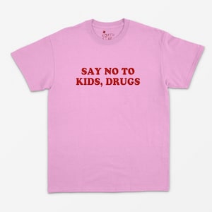 May include: A pale pink t-shirt featuring the red text "SAY NO TO KIDS, DRUGS". The shirt has a crew neck and short sleeves. The brand logo "NORTH STAR" is printed in white on the upper left.