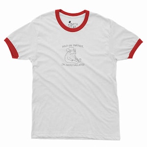 May include: White ringer t-shirt with red trim on the collar and sleeves. The shirt features a cartoon frog wearing a hat, with the text "HOLD ON PARTNER, I'M OVERSTIMULATED". The North Star logo is on the collar.