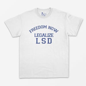 May include: White t-shirt featuring the blue text "FREEDOM NOW LEGALIZE LSD". The text is arranged in an arc above the word "LEGALIZE" and stacked above "LSD". The shirt has a crew neck and short sleeves. A small logo is visible at the top.