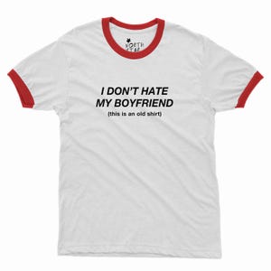 May include: White ringer t-shirt with red trim on the collar and sleeves. The shirt has the text "I DON'T HATE MY BOYFRIEND (this is an old shirt)" in black. A small logo is at the top.