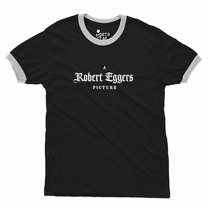 May include: A black ringer t-shirt with light gray trim on the collar and sleeves. The chest features white text that reads A Robert Eggers Picture in a gothic font. The inner neck label displays the text North Star with a small star icon.
