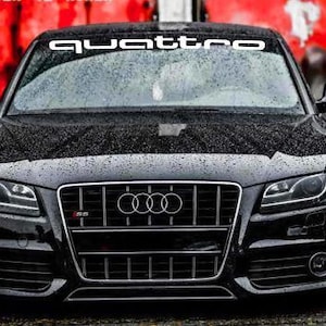 Op de afbeelding: Een zwarte Audi S5 coupé met het woord "quattro" in het wit over de bovenkant van de voorruit. De auto heeft een zwarte grille met het Audi-logo en het S5-embleem. De achtergrond is een vage rode en grijze kleur.