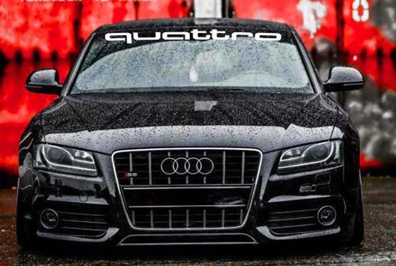 May include: A black Audi S5 coupe with the word "quattro" in white across the top of the windscreen. The car has a black grill with the Audi logo and the S5 emblem. The background is a blurred red and grey.