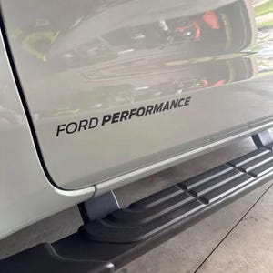 Könnte beinhalten: Ein Ausschnitt der Seitenwand eines weißen Fahrzeugs mit den Worten "FORD PERFORMANCE" in schwarzer Schrift. Darunter befindet sich ein schwarzes Trittbrett mit strukturierter Oberfläche.