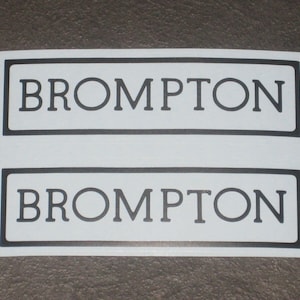 May include: Two white rectangular stickers with the word "BROMPTON" in black capital letters. Each sticker has a black border. The stickers are on a gray surface.