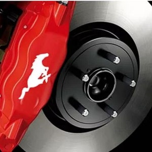 Könnte beinhalten: Nahaufnahme eines roten Ferrari-Bremssattels mit dem weißen Logo des springenden Pferdes. Der Bremssattel ist mit einer silbernen Bremsscheibe und einer schwarzen Nabe vor dunklem Hintergrund kombiniert.