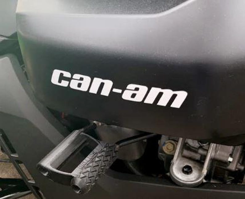 Puede incluir: Primer plano de un componente negro de veh&iacute;culo con el texto blanco "can-am". Debajo, se observa un reposapi&eacute;s negro con superficie texturizada. La imagen destaca la marca y el dise&ntilde;o del veh&iacute;culo.
