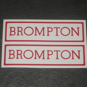 May include: Two rectangular stickers with the word "BROMPTON" in red capital letters. The text is inside a white rectangle with a thin red border. The stickers are on a dark gray surface.