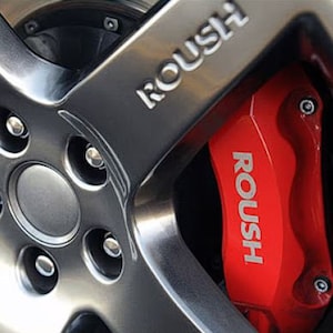 Puede incluir: Primer plano de una llanta de coche con acabado gris plateado. La llanta presenta el nombre de la marca "ROUSH" en relieve. Una pinza de freno roja brillante con el logotipo "ROUSH" es visible detrás de los radios de la llanta.
