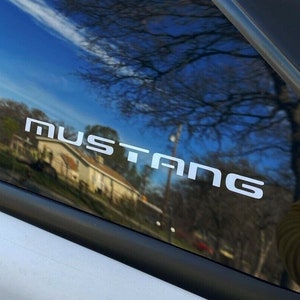 May include: White "MUSTANG" decal on a car window. The decal is in a bold, sans-serif font. The window reflects a blue sky, trees, and a house.