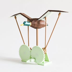 May include: A wooden duck toy with brown wings and body, a blue and green head, and a yellow beak. The duck is suspended above two light green gears. The gears are connected to a wooden base with wooden supports.