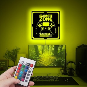 Może przedstawiać: Czarny i żółty znak świetlny LED Gamer Zone z grafiką kontrolera i napisem "LOADING...". Pilot zdalnego sterowania z wieloma opcjami kolorów jest trzymany przed znakiem. Znak jest zamontowany na żółtej ścianie nad konfiguracją komputera.