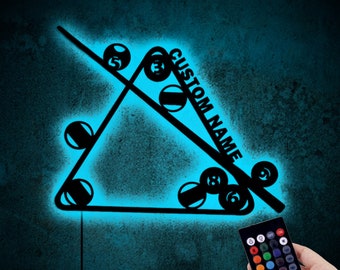 May include: A black and blue LED wall art piece featuring a pool table design. The design includes a triangle of pool balls, a cue stick, and the text "CUSTOM NAME". A remote control is held in the hand.