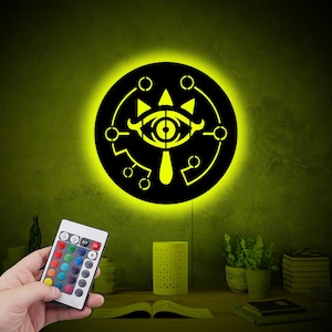 Puede incluir: Un aplique de pared negro circular con un recorte del símbolo del ojo Sheikah iluminado en amarillo. La luz emite un brillo amarillo intenso y se controla mediante un mando a distancia con varios botones de colores para ajustar la iluminación.