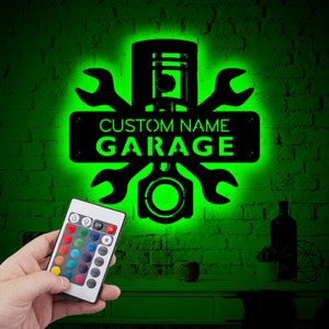 Peut inclure: Une enseigne en métal noir avec un design de piston et de clé à molette. L'enseigne indique "CUSTOM NAME GARAGE" et est éclairée d'une lumière verte. Une télécommande avec des lumières LED changeantes de couleur est au premier plan.