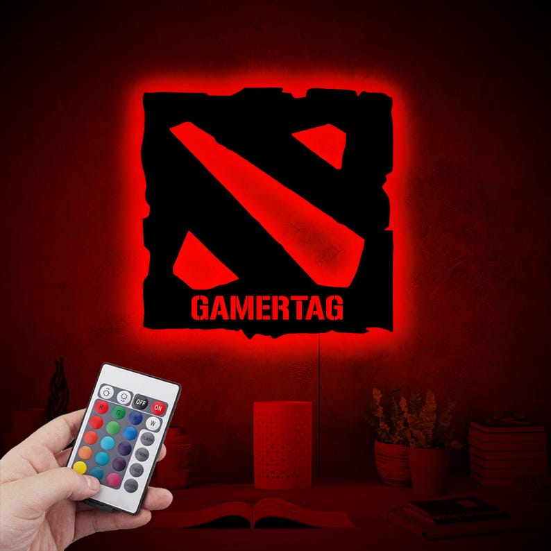 May include: A red and black gamertag light with a remote control. The light features the Dota 2 logo and the word "GAMERTAG".