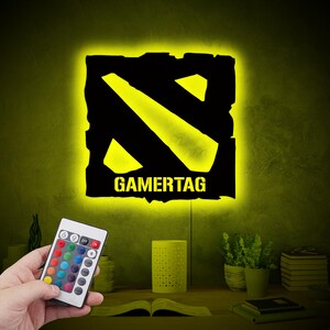 May include: A black and yellow gamertag light with a remote control. The light is shaped like the Dota 2 logo and has the word "GAMERTAG" written below it.