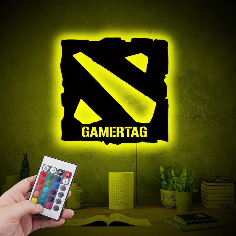 May include: A black and yellow gamertag light with a remote control. The light is shaped like the Dota 2 logo and has the word "GAMERTAG" written below it.