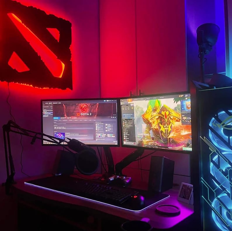 May include: A gaming setup with two monitors, a keyboard, and a mouse. The monitors are displaying a game of Dota 2 and a game of Sand King. The setup is lit with red and blue lights. The Dota 2 logo is displayed on the wall behind the monitors.