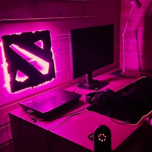 Puede incluir: Una configuración de juego iluminada en rosa con una computadora portátil, un monitor, una alfombrilla de ratón y una luz con el logotipo de Dota 2.