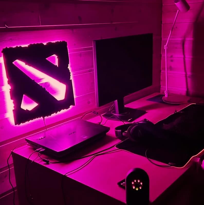 May include: A pink-lit gaming setup with a laptop, a monitor, a mousepad, and a Dota 2 logo light.