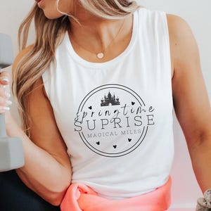 May include: A woman wearing a white tank top with the text "Springtime Surprise Magical Miles" in a circle with a castle graphic. She is holding a gray dumbbell and wearing black leggings and pink sweatpants.