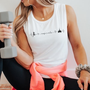 May include: White sleeveless tank top with a black graphic of a running person and a castle with the text "magic x miles" on a white background. The person is wearing black leggings and white sneakers and is holding a gray dumbbell.