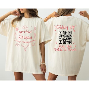 May include: Two white t-shirts with pink text and graphics. The front shirt says "gettin' hitched" with a cactus, horseshoe, cowboy hat, and other western-themed graphics. The back shirt says "Giddy Up" with a QR code and "Buy the Bride a Drink".
