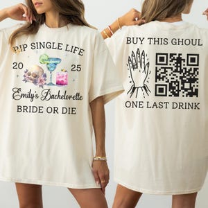 May include: Cream-colored t-shirt with front graphic: "PIP SINGLE LIFE 20 25 Emily's Bachelorette BRIDE OR DIE" with skull, cocktails, and floral elements. Back graphic: "BUY THIS GHOUL ONE LAST DRINK" with hand, ring, and QR code.