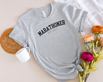 May include: Gray t-shirt with black text that reads "MARATHONER EST 2023".