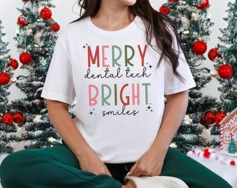 May include: White t-shirt with the text "Merry dental tech bright smiles" in colorful letters. The shirt is worn by a person sitting in front of a Christmas tree.