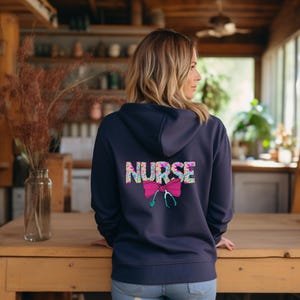 May include: A navy blue hoodie with a floral pattern "NURSE" design on the back. The design includes a pink bow and a stethoscope.