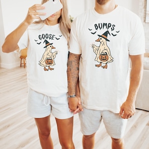 May include: Two matching white t-shirts with a Halloween goose design. The goose wears a witch hat and carries a pumpkin. One shirt says "Goose" and the other says "Bumps". Both shirts have bats above the goose.
