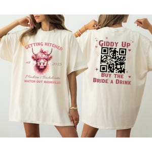 May include: Two white t-shirts with a pink and black design. The front shirt has a cartoon cow with sunglasses and a bow. The text reads "Getting Hitched est 2025 Madison's Bachelorette Watch Out Nashville!" The back shirt has a QR code and the text "Giddy Up Buy the Bride a Drink".