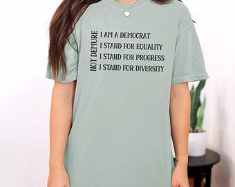 May include: A light green t-shirt with the text "NOT DEMURE" printed vertically on the left side and the text "I AM A DEMOCRAT I STAND FOR EQUALITY I STAND FOR PROGRESS I STAND FOR DIVERSITY" printed horizontally on the right side.